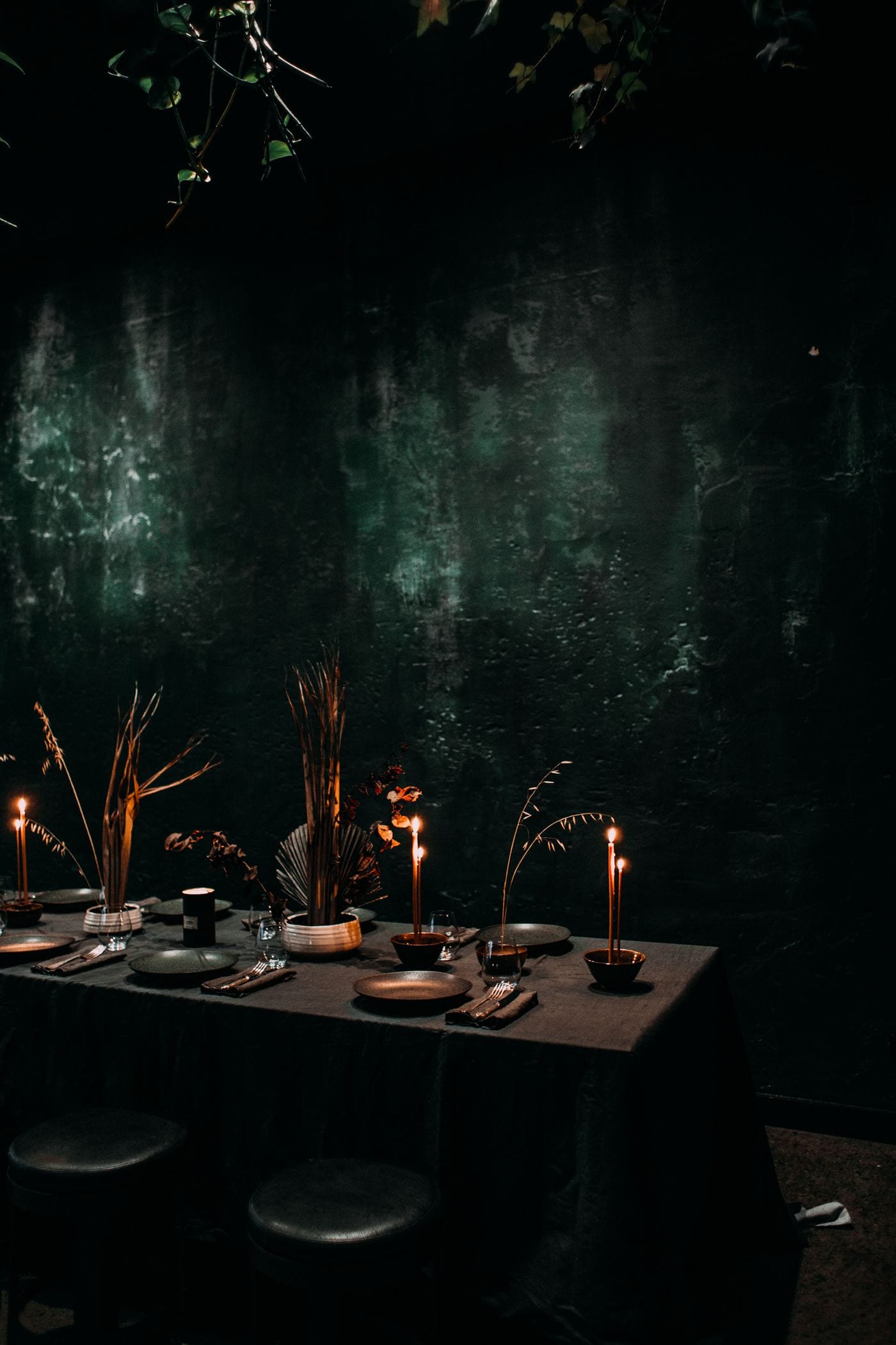 Long table of dried stems and lit tapers against a deep green wall